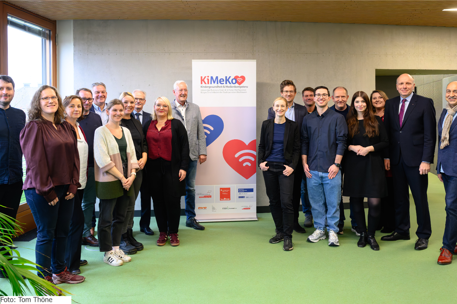 Eine Gruppe von Menschen steht neben einem Aufsteller mit der Aufschrift KiMeKo.