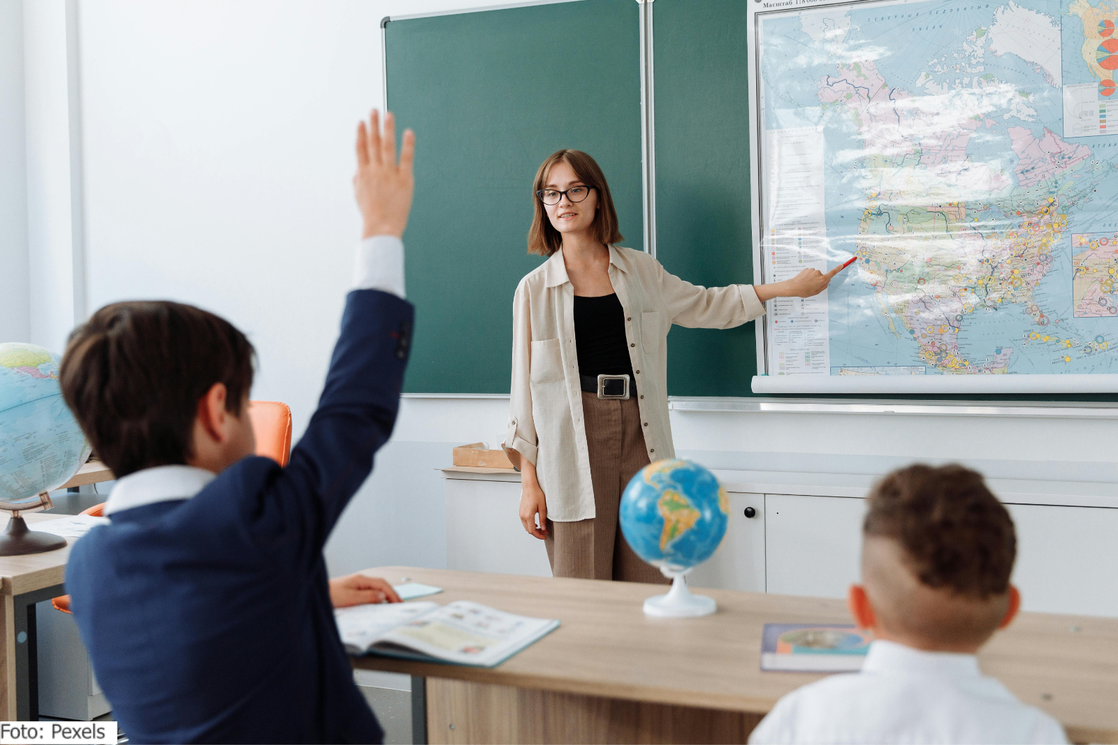 Eine junge Lehrerin zeigt auf eine Landkarte, zwei Schüler melden sich